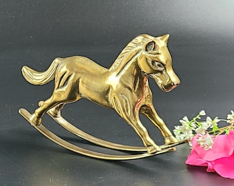 Puede incluir: Una figurita de caballo de balancín de latón de color dorado. El caballo está de pie sobre una base de balancín con dos soportes curvos.
