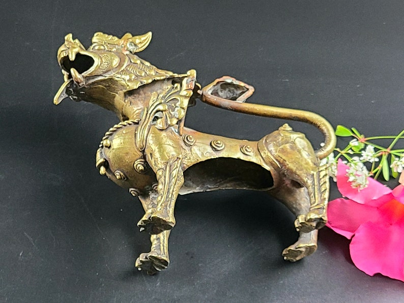Puede incluir: Una escultura de metal dorado de un le&oacute;n m&iacute;tico con detalles intrincados. El le&oacute;n est&aacute; de pie sobre sus cuatro patas con la cabeza girada hacia un lado y la boca abierta. La escultura mide aproximadamente 15 cm de alto.