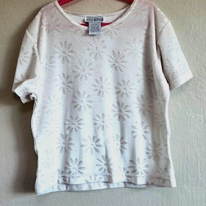 May include: A white short-sleeved shirt with a daisy pattern. The shirt has a scoop neck and is hanging on a pink hanger. The tag reads "ADELE NORMAN".