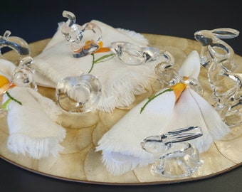 Könnte beinhalten: Satz mit sechs klaren Glas-Serviettenringen, jeder in Form eines Tieres. Die Ringe sind auf einer weißen Serviette mit Blumenmuster und einer beigefarbenen, strukturierten Oberfläche angeordnet. Zu den Tieren gehören ein Elefant, ein Kaninchen und ein Apfel.