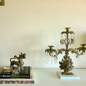 Puede incluir: Un candelabro dorado con prismas de cristal y una base de m&aacute;rmol. El candelabro tiene tres brazos y est&aacute; decorado con motivos florales. Una figurita dorada de una criatura m&iacute;tica est&aacute; sobre una pila de libros con el texto "The Broker" y "John Grisham" visible.