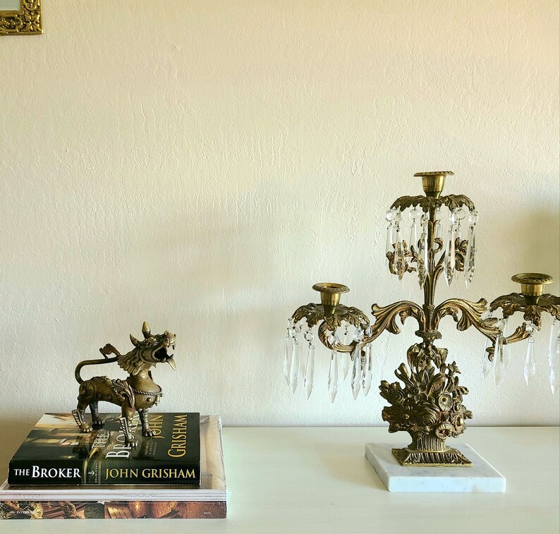 Puede incluir: Un candelabro dorado con prismas de cristal y una base de m&aacute;rmol. El candelabro tiene tres brazos y est&aacute; decorado con motivos florales. Una figurita dorada de una criatura m&iacute;tica est&aacute; sobre una pila de libros con el texto "The Broker" y "John Grisham" visible.