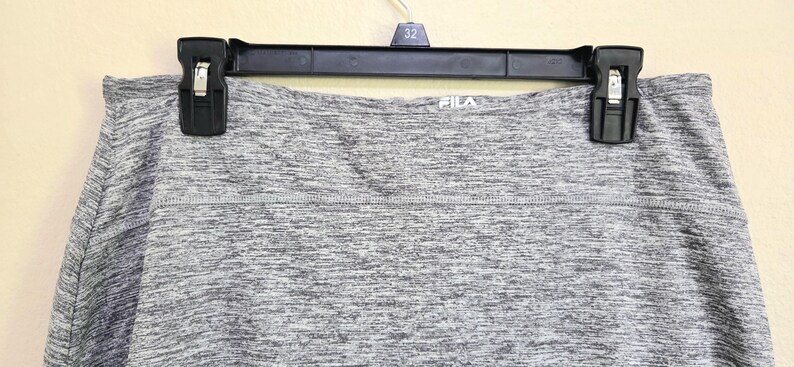May include: Gray athletic skirt hanging on a black hanger. The skirt has a heathered pattern and the brand name "FILA" is visible. The skirt is made of a stretchy material, suitable for sports and exercise.