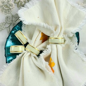 Può includere: Anelli per tovaglioli color crema con finiture dorate sono esposti con tovaglioli bianchi con frange. Gli anelli per tovaglioli sono disposti su un piatto di vetro verde acqua. I tovaglioli sono piegati e decorati con gli anelli.