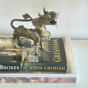 Puede incluir: Una estatua de metal dorado de un le&oacute;n m&iacute;tico con detalles intrincados y una cola levantada. La estatua est&aacute; sentada sobre dos libros, uno titulado "The Broker" de John Grisham.