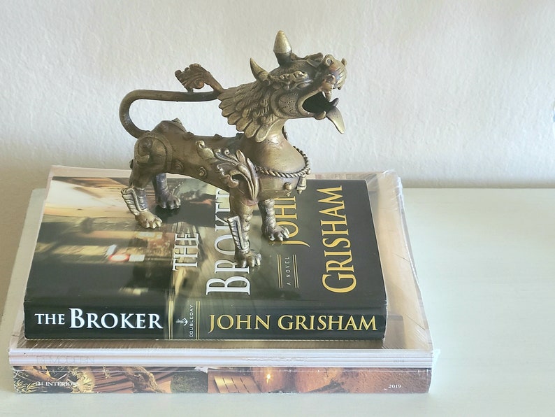 Puede incluir: Una estatua de metal dorado de un le&oacute;n m&iacute;tico con detalles intrincados y una cola levantada. La estatua est&aacute; sentada sobre dos libros, uno titulado "The Broker" de John Grisham.