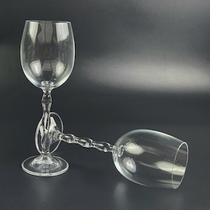 May include: Two clear wine glasses with a twisted stem design. One glass is upright, the other is lying on its side. The glasses are made of transparent glass and are set against a dark background.
