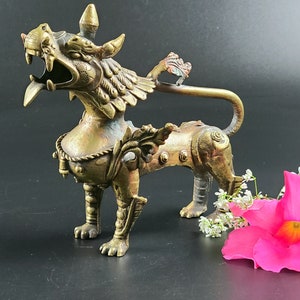 Puede incluir: Una escultura de metal dorado de una criatura m&iacute;tica con alas y una cola larga. La criatura tiene una expresi&oacute;n feroz y est&aacute; decorada con detalles intrincados.