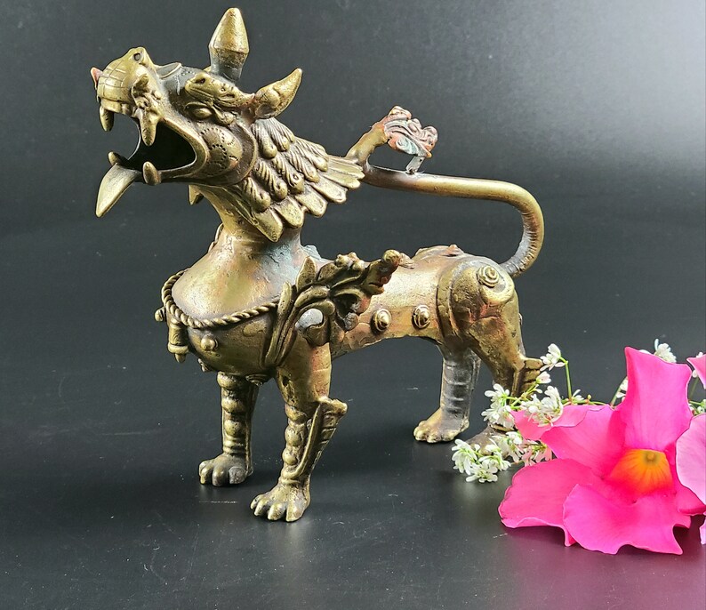 Puede incluir: Una escultura de metal dorado de una criatura m&iacute;tica con alas y una cola larga. La criatura tiene una expresi&oacute;n feroz y est&aacute; decorada con detalles intrincados.