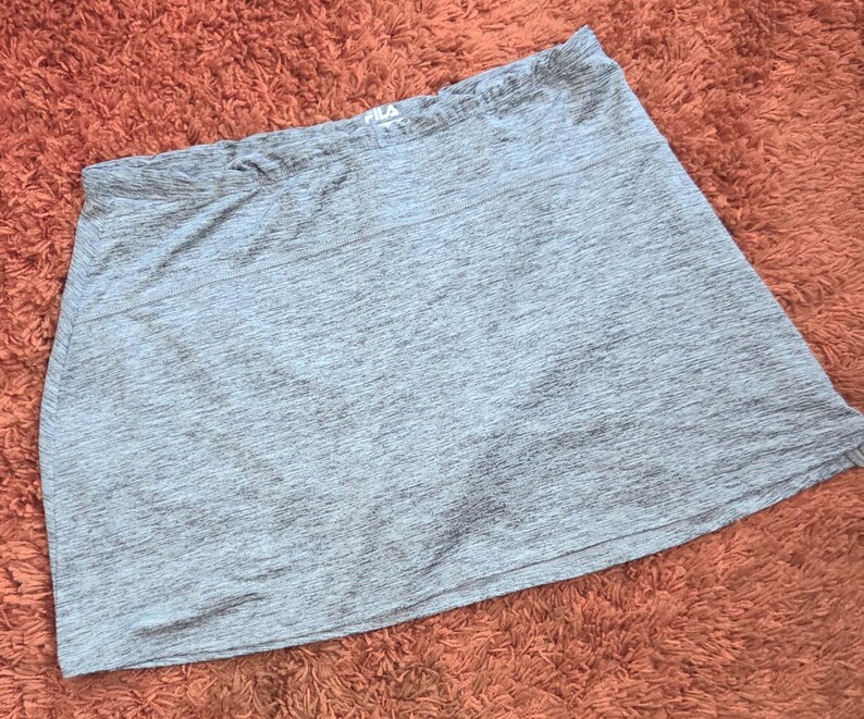 May include: A gray athletic skirt with a slightly flared design. The skirt has a comfortable waistband and a subtle textured pattern. The Fila logo is visible on the waistband. This activewear skirt is suitable for various sports and activities.