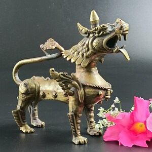 Puede incluir: Una escultura de metal dorado de una criatura m&iacute;tica con alas y una cola larga. La criatura tiene una expresi&oacute;n feroz y est&aacute; de pie sobre sus cuatro patas. La escultura est&aacute; detallada con intrincados tallados y es una obra de arte &uacute;nica.