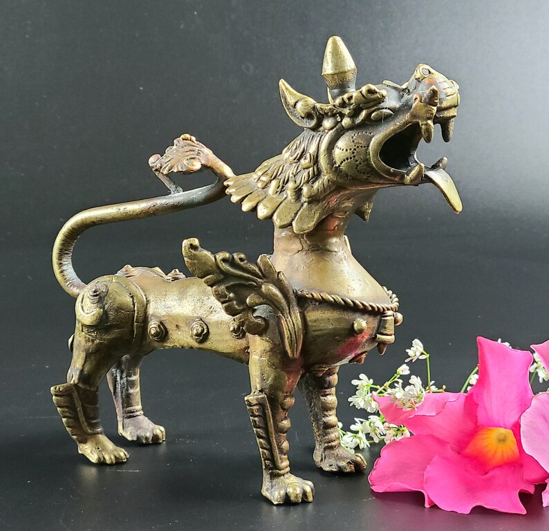 Puede incluir: Una escultura de metal dorado de una criatura m&iacute;tica con alas y una cola larga. La criatura tiene una expresi&oacute;n feroz y est&aacute; de pie sobre sus cuatro patas. La escultura est&aacute; detallada con intrincados tallados y es una obra de arte &uacute;nica.