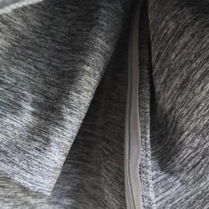 May include: Close-up of gray athletic fabric with a zipper. The fabric has a heathered texture, with shades of gray creating a subtle pattern. The zipper is a matching gray color and runs vertically.