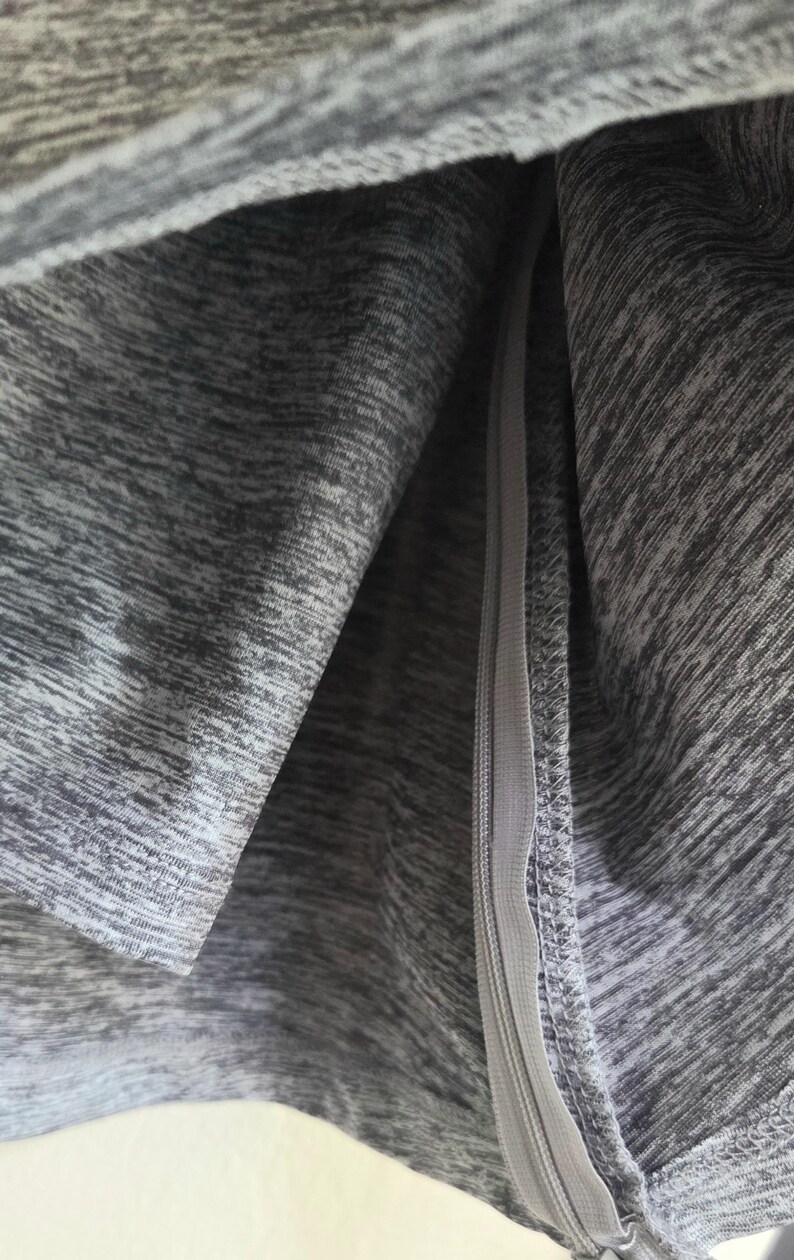 May include: Close-up of gray athletic fabric with a zipper. The fabric has a heathered texture, with shades of gray creating a subtle pattern. The zipper is a matching gray color and runs vertically.