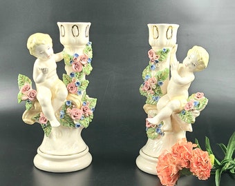 May include: Two white porcelain candle holders featuring cherub figures and floral accents. Each holder depicts a cherub with wings, climbing a floral stem with pink, white and blue flowers.