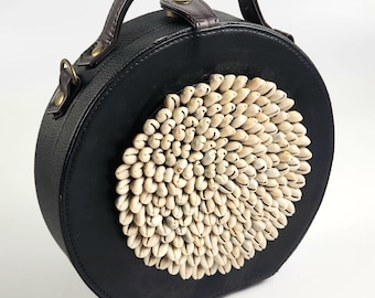 May include: A black round handbag with a brown leather handle and a circular design of white seashells.
