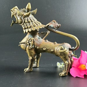 Puede incluir: Una escultura de metal dorado de un le&oacute;n m&iacute;tico con detalles intrincados y una cola enroscada. La escultura est&aacute; sobre una superficie negra con flores rosas en primer plano.