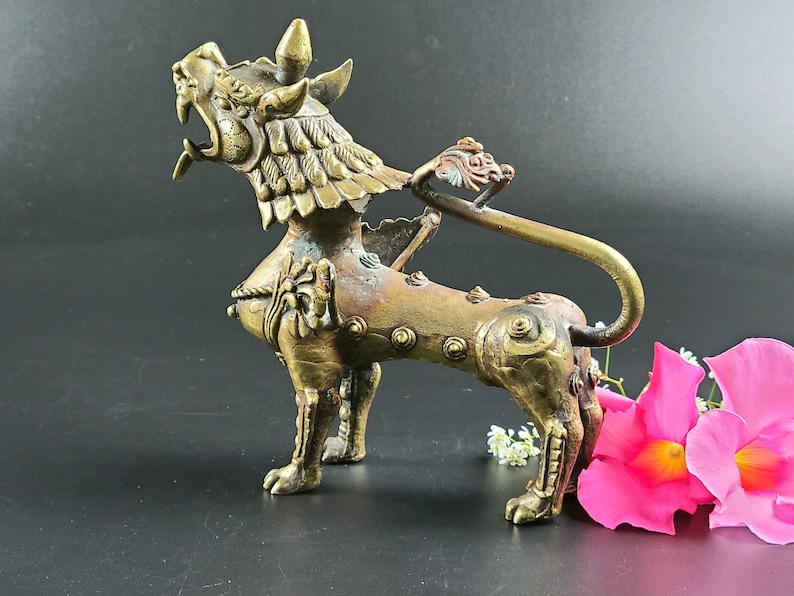 Puede incluir: Una escultura de metal dorado de un le&oacute;n m&iacute;tico con detalles intrincados y una cola enroscada. La escultura est&aacute; sobre una superficie negra con flores rosas en primer plano.