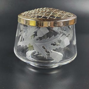 May include: A clear glass vase with etched leaf designs. The vase has a silver rim and a metal grid lid. The design is a classic and elegant piece for floral arrangements. The vase is a decorative item.