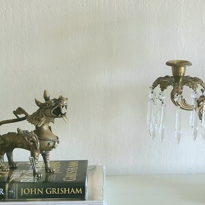 Puede incluir: Una estatua de metal dorado de una criatura m&iacute;tica con una cola larga y garras afiladas, sentada sobre un libro titulado "The Broker" de John Grisham. La estatua est&aacute; junto a un candelabro de metal dorado con prismas de cristal.