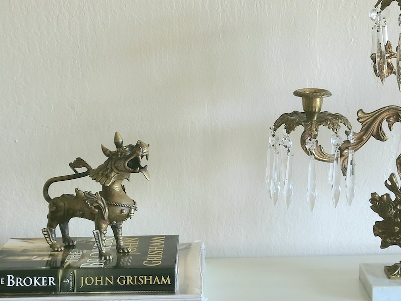 Puede incluir: Una estatua de metal dorado de una criatura m&iacute;tica con una cola larga y garras afiladas, sentada sobre un libro titulado "The Broker" de John Grisham. La estatua est&aacute; junto a un candelabro de metal dorado con prismas de cristal.
