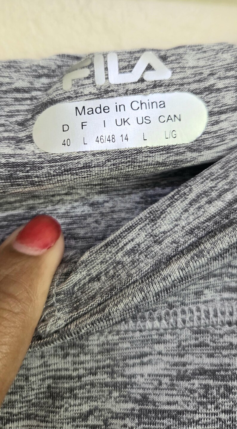May include: Close-up of a gray and white athletic top with the Fila logo. The tag reads "Made in China" with size information. The fabric has a textured, heathered appearance.