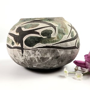 May include: A round, gray ceramic vase with a black and green abstract design. The vase has a wide mouth and a rounded bottom.
