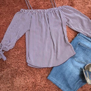 Könnte beinhalten: Ein gestreiftes Off-Shoulder-Top mit langen Ärmeln und Bindedetails, kombiniert mit einer blauen Jeans. Das Oberteil hat dünne Träger und ein vertikales Streifenmuster in Weiß und Dunkelviolett. Silberne Absätze sind sichtbar.