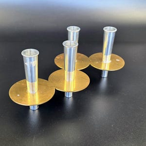 Peut inclure: Quatre bougeoirs vintage. Chaque bougeoir a un sommet cylindrique argenté et une base circulaire dorée. Les bougeoirs sont disposés sur une surface sombre, mettant en valeur leur design et leurs finitions métalliques.