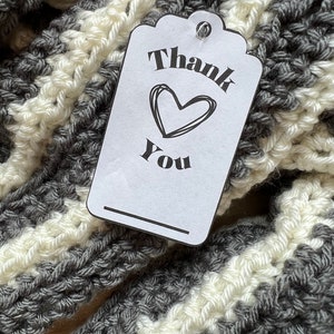 May include: A white gift tag with black text that says "Thank You" with a heart drawn above the word "You". The tag is attached to a gray and white striped crocheted scarf.