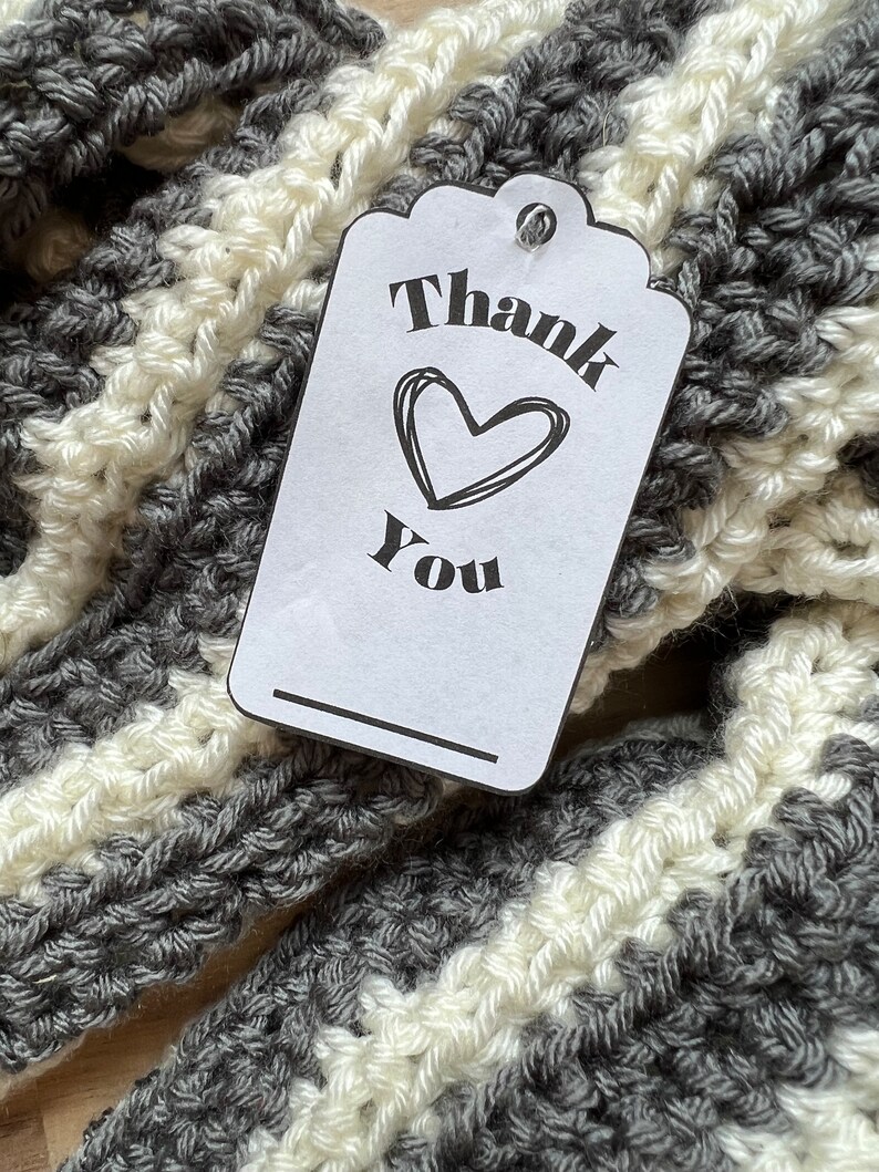 May include: A white gift tag with black text that says "Thank You" with a heart drawn above the word "You". The tag is attached to a gray and white striped crocheted scarf.