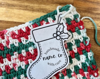 May include: A white crocheted dishcloth with red and green stripes. A paper tag with a stocking shape is attached to the dishcloth. The tag has a holly design and the text "handmade name. co with care".