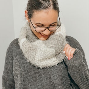 May include: A woman wearing a gray sweater with a white and gray fuzzy scarf. She is wearing glasses and smiling.