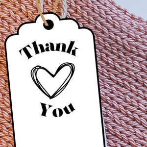 May include: A white gift tag with black text that says "Thank You" with a heart drawn in the middle. The tag is on a pink and orange woven background.