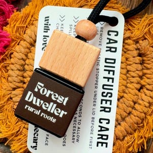 Peut inclure: Diffuseur de voiture avec un dessus en bois et une base marron, avec le texte "Forest Dweller rural roots". Le produit est attaché à une étiquette blanche avec des instructions d'entretien et un cordon noir. Le fond est une surface orange texturée.