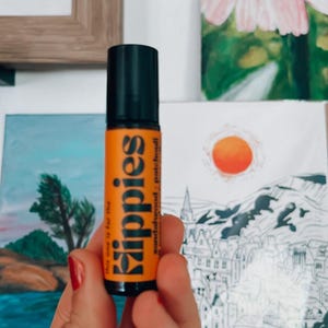 Peut inclure: Flacon de roll-on d'huile essentielle orange et noir avec le texte "Kippies" et "sandalwood - patchouli". Le flacon est tenu devant une peinture de fleur et un dessin de paysage.