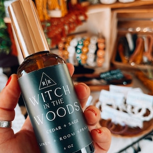 May include: A 4 ounce glass bottle of room spray with a black label that reads "Witch in the Woods Cedar + Sage Linen + Room Spray". The bottle has a gold cap and is held in a hand.