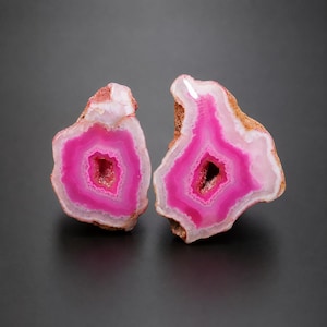 Könnte beinhalten: Zwei polierte Achat-Scheiben mit leuchtend pinkfarbenen Zentren, umgeben von konzentrischen Ringen in hellem Rosa und Weiß. Die Außenkanten dieser Mineralien sind rau, strukturiert und braun und zeigen eine natürliche kristalline Beschaffenheit.
