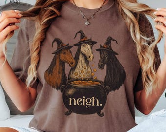 May include: Espresso-colored t-shirt featuring a whimsical design of three horses wearing witch hats, gathered around a bubbling cauldron. The cauldron has the word "neigh" printed on it. The shirt is a comfortable, casual style.
