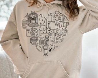 Puede incluir: Una sudadera con capucha beige con un contorno negro de un corazón lleno de diversos equipos de fotografía, incluyendo cámaras, lentes, un trípode, un flash y una bombilla.