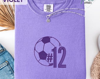 May include: A violet Comfort Colors t-shirt with a soccer ball graphic and the number 12. A silver necklace with a flower pendant is draped across the shirt. The word "VIOLET" is in the upper left corner.