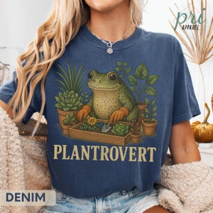 May include: Denim blue t-shirt featuring a frog tending plants in a garden setting. The frog is surrounded by potted plants and a small garden box with flowers and gardening tools. The word "PLANTROVERT" is printed in a bold font.
