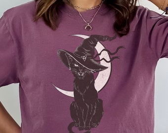 May include: A purple t-shirt with a black cat wearing a witch's hat and sitting on a crescent moon. The cat has a mischievous grin and a triangular symbol on its hat.