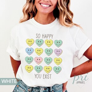 May include: White t-shirt with a graphic of colorful heart-shaped smiley faces and the text "So Happy You Exist".