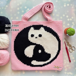 Puede incluir: Una bolsa de mano de crochet rosa con un diseño yin yang en blanco y negro con dos gatos. La bolsa tiene una correa larga y está hecha con un punto de crochet sencillo.
