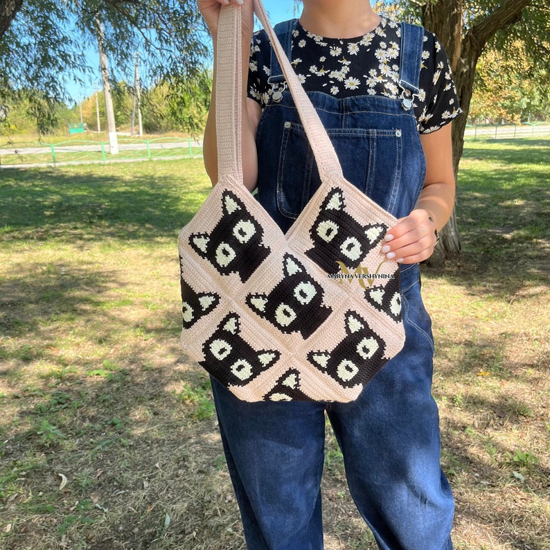 May include: A pink crocheted tote bag with black and white cat faces. The bag has a long strap and is being held by a person.
