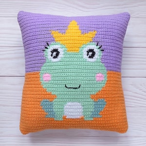 May include: A crocheted pillow with a frog wearing a crown. The frog is green with white eyes and pink cheeks. The pillow is purple and orange.