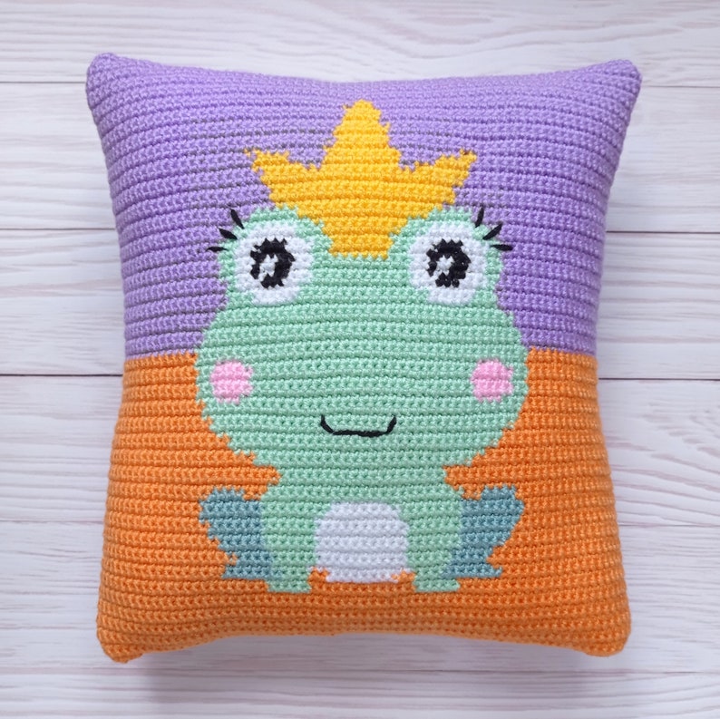 May include: A crocheted pillow with a frog wearing a crown. The frog is green with white eyes and pink cheeks. The pillow is purple and orange.