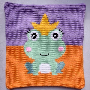 May include: A crocheted pillow with a purple and orange background featuring a green frog wearing a yellow crown. The frog has white eyes, a pink nose, and a white belly.