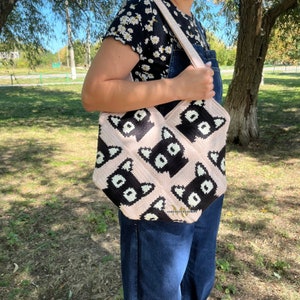 May include: A pink and black crocheted tote bag with a cat face pattern. The bag has a long strap and is being held by a person.